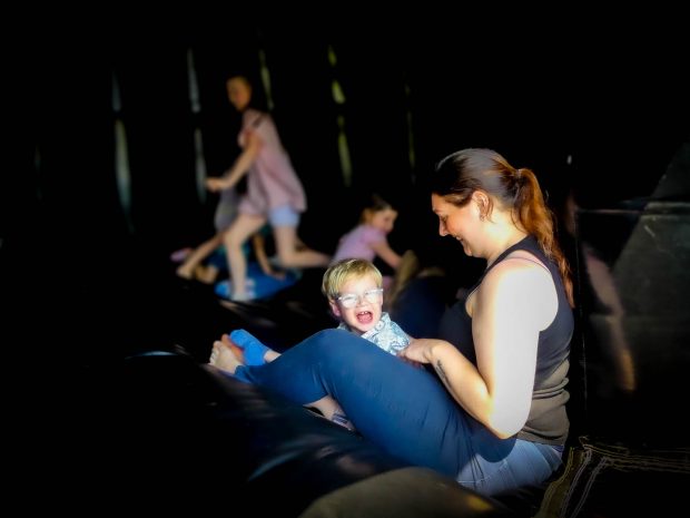 A smiling woman and a happy child sit together in an indoor play area with other children playing in the background. Cara’s Story