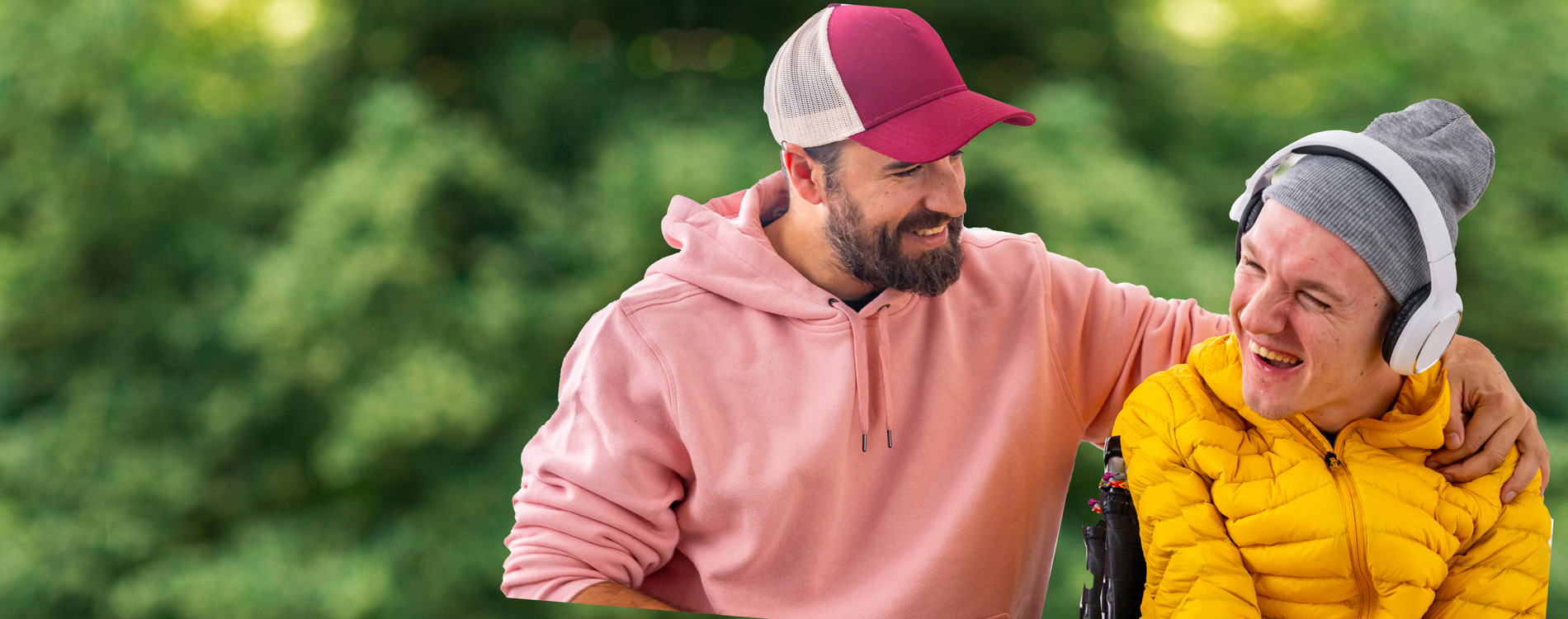 Two men outdoors, one in a pink hoodie and cap, the other in a wheelchair wearing headphones and a yellow jacket.