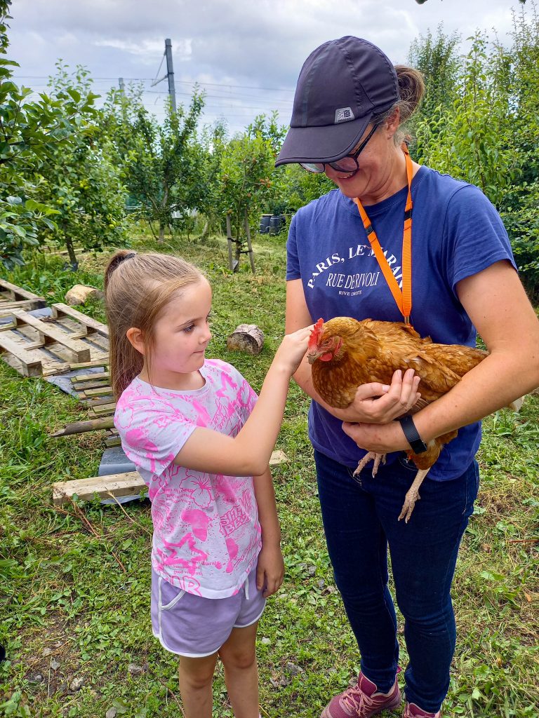 A young girl pets a chicken held by a smiling woman outdoors in a grassy area with trees.