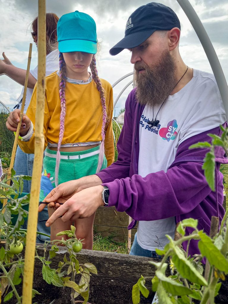A man shows a girl with purple braids how to tend tomato plants in a garden.