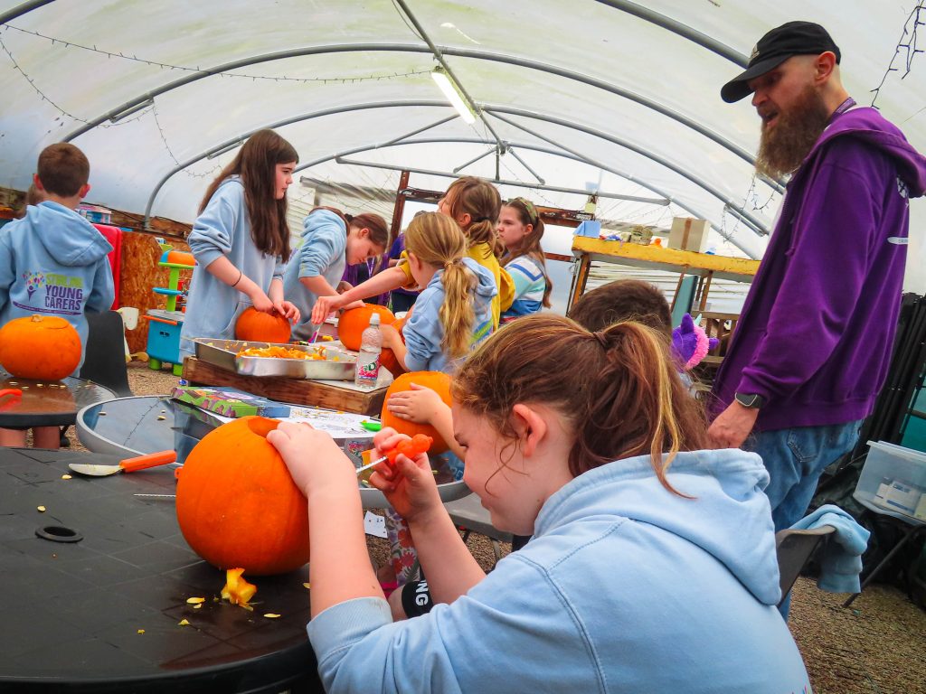Children carve pumpkins at a table in a greenhouse while an adult supervises.
