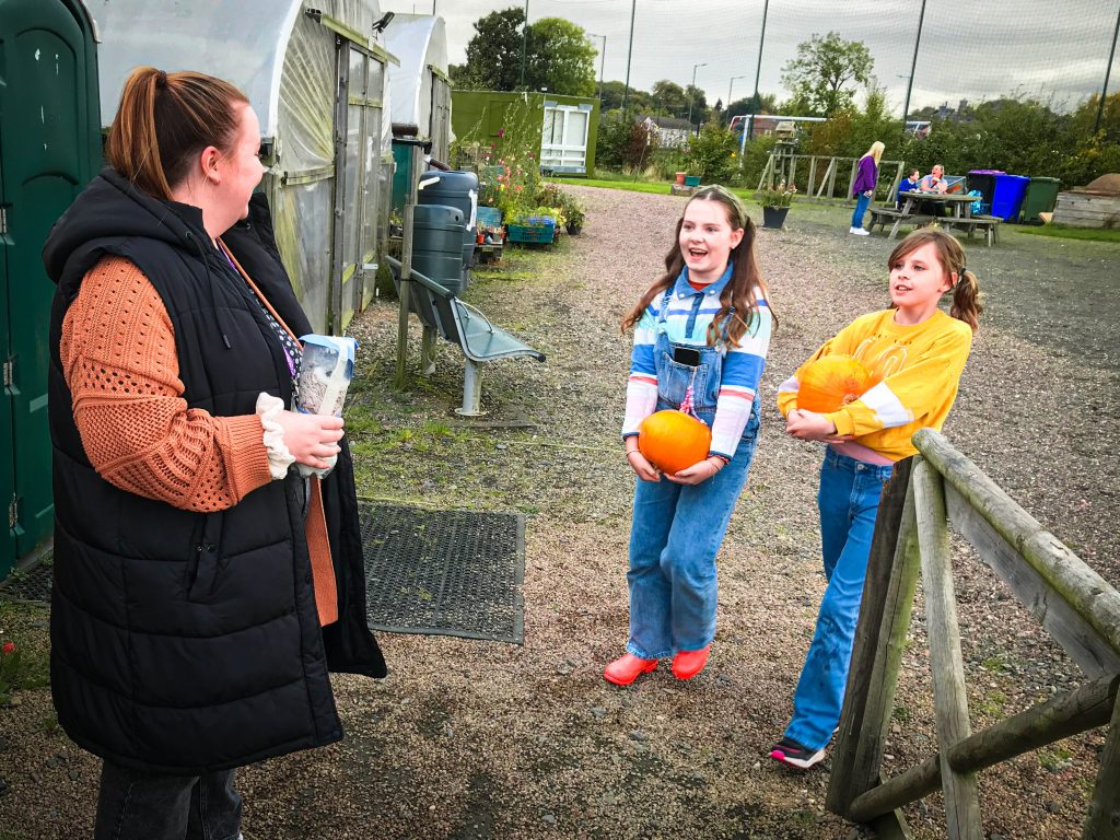 A woman talks to two girls holding pumpkins beside a garden with raised beds on a cloudy day.