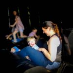 A smiling woman sits with a laughing young boy in a dark play area while other children play in the background.