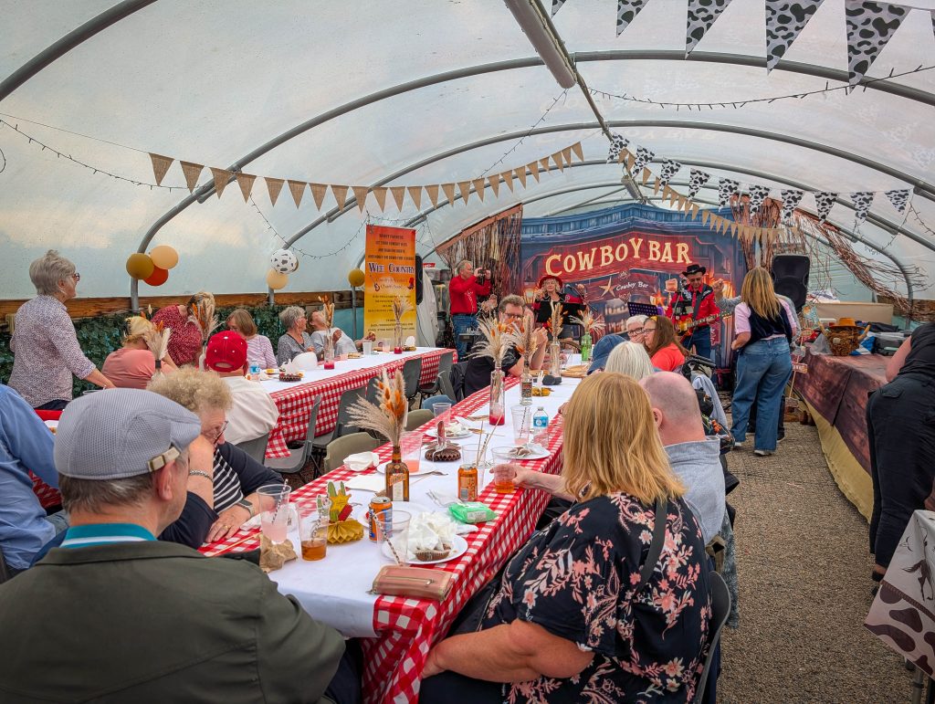 People sit at long tables covered with red checkered cloths, watching a band perform on a stage with a sign reading Cowboy Bar.