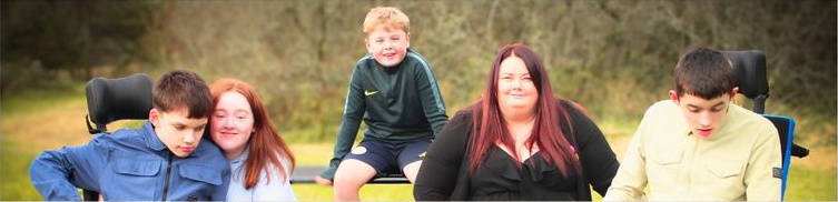 A caucasian family sit together. In the centre is a young, blonde-haired boy sat on a bench. In front is his mother dressed in black with long dark red hair.  To the right is a boy in an electric wheelchair wearing a blue top being hugged by a girl with ginger hair wearing a light blue hoodie. To the right is a young adult male in a yellow top sitting in an electric wheelchair. Both boys in wheelchairs have complex disabilities.