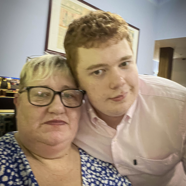 A woman wearing glasses and a man in a pink shirt pose closely together for a selfie, both with subtle smiles.