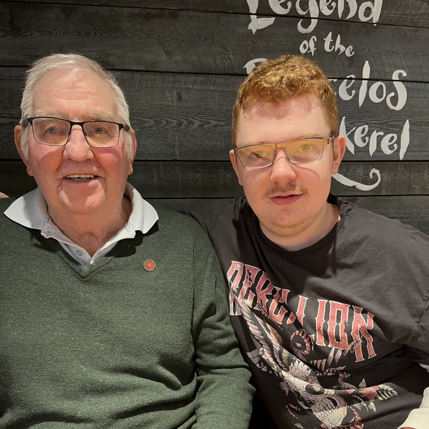 Two men sit side by side, smiling, in front of a dark wooden wall with white writing.