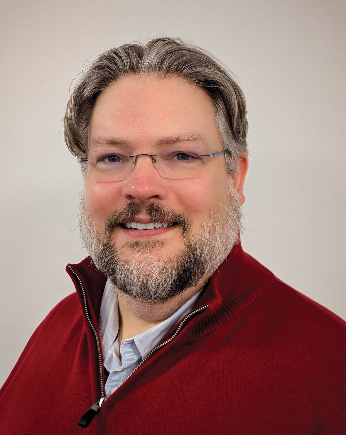 A man with glasses, gray hair, and a beard wears a red sweater and smiles in front of a light background.