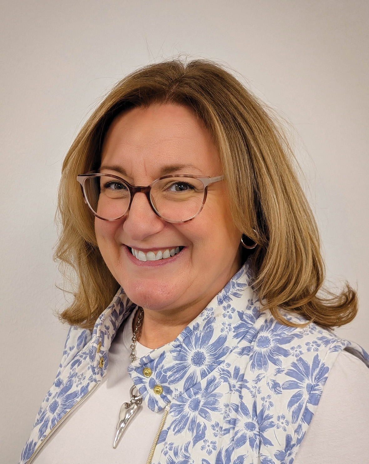A woman with blonde hair and glasses, smiling and dressed in a floral vest with a necklace, stands in front of a plain background.