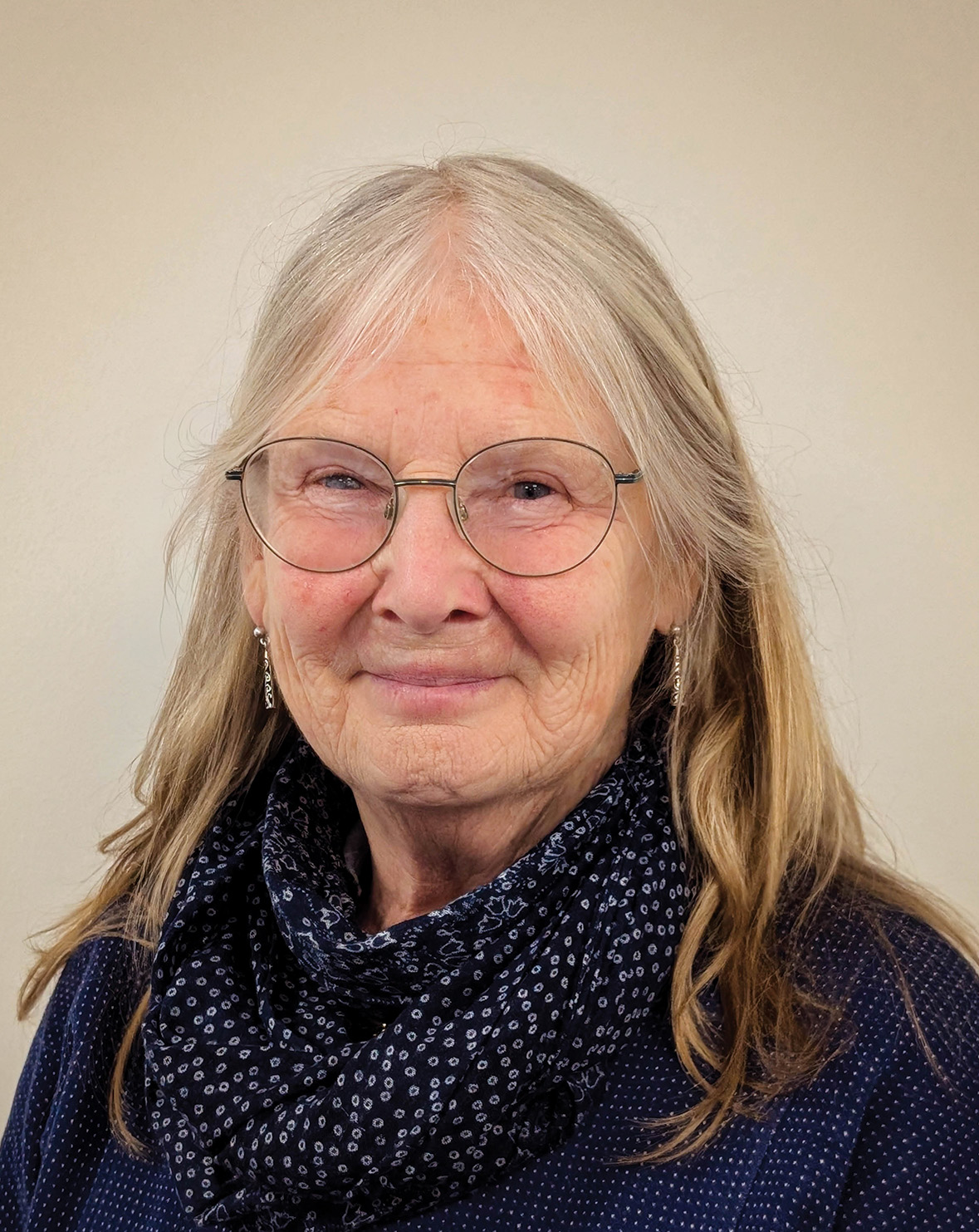 Smiling elderly woman with long gray hair, wearing glasses and a blue scarf, in front of a plain background.
