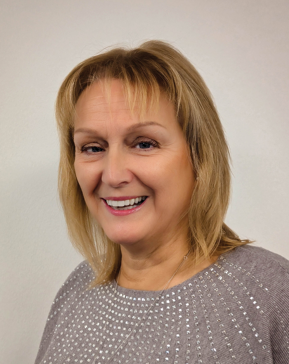 Middle-aged woman with shoulder-length blonde hair smiles, wearing a light gray sweater with dots.