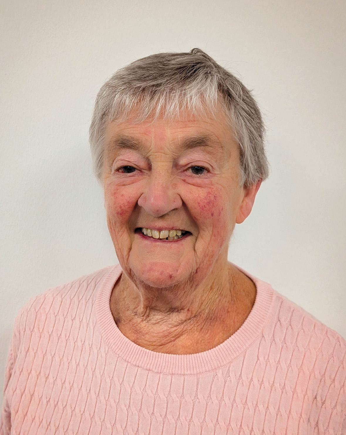 An elderly woman with short gray hair and a light pink sweater smiles while standing against a plain background.