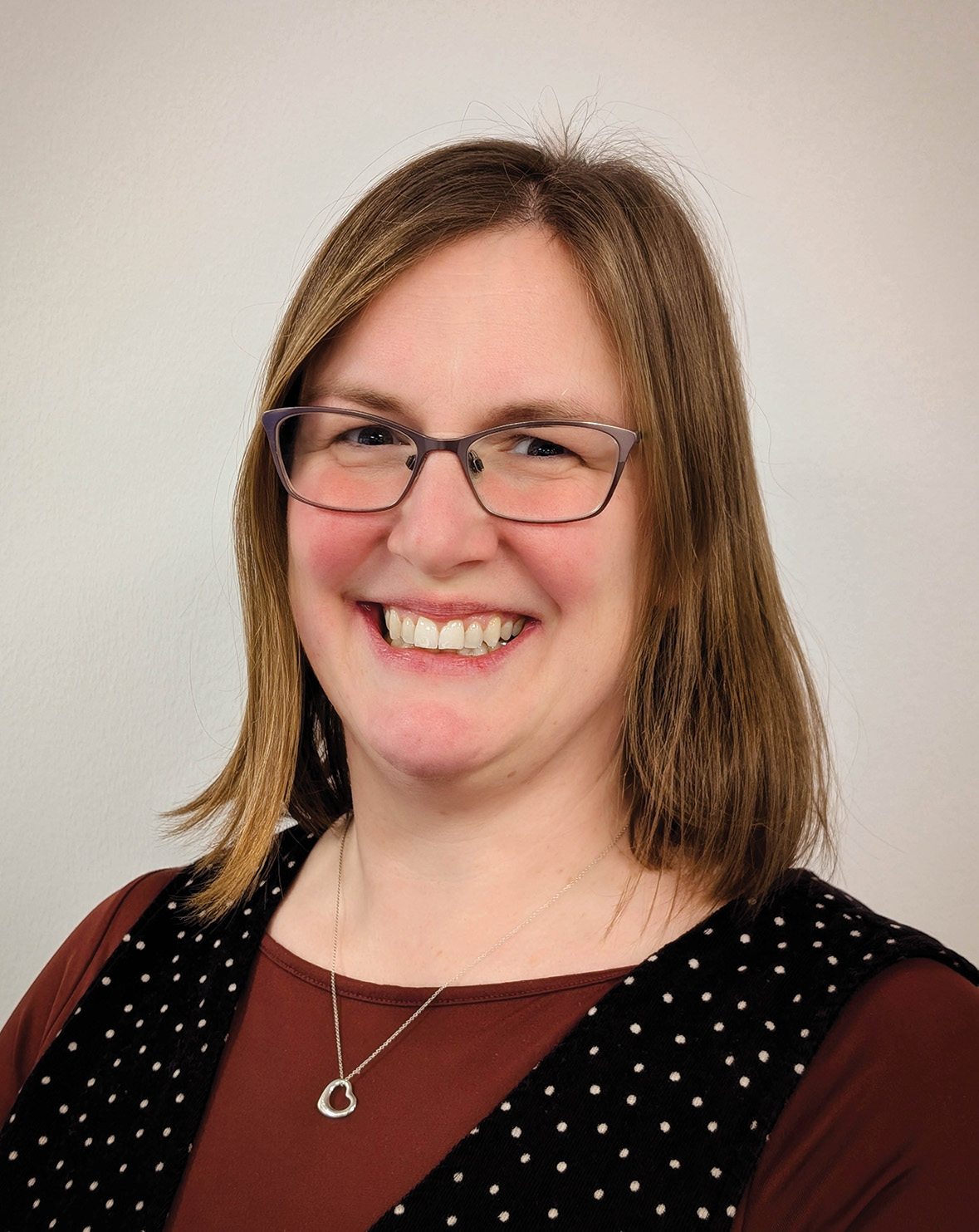 A woman with shoulder-length hair, wearing glasses, a necklace, and a polka dot vest, smiles at the camera.