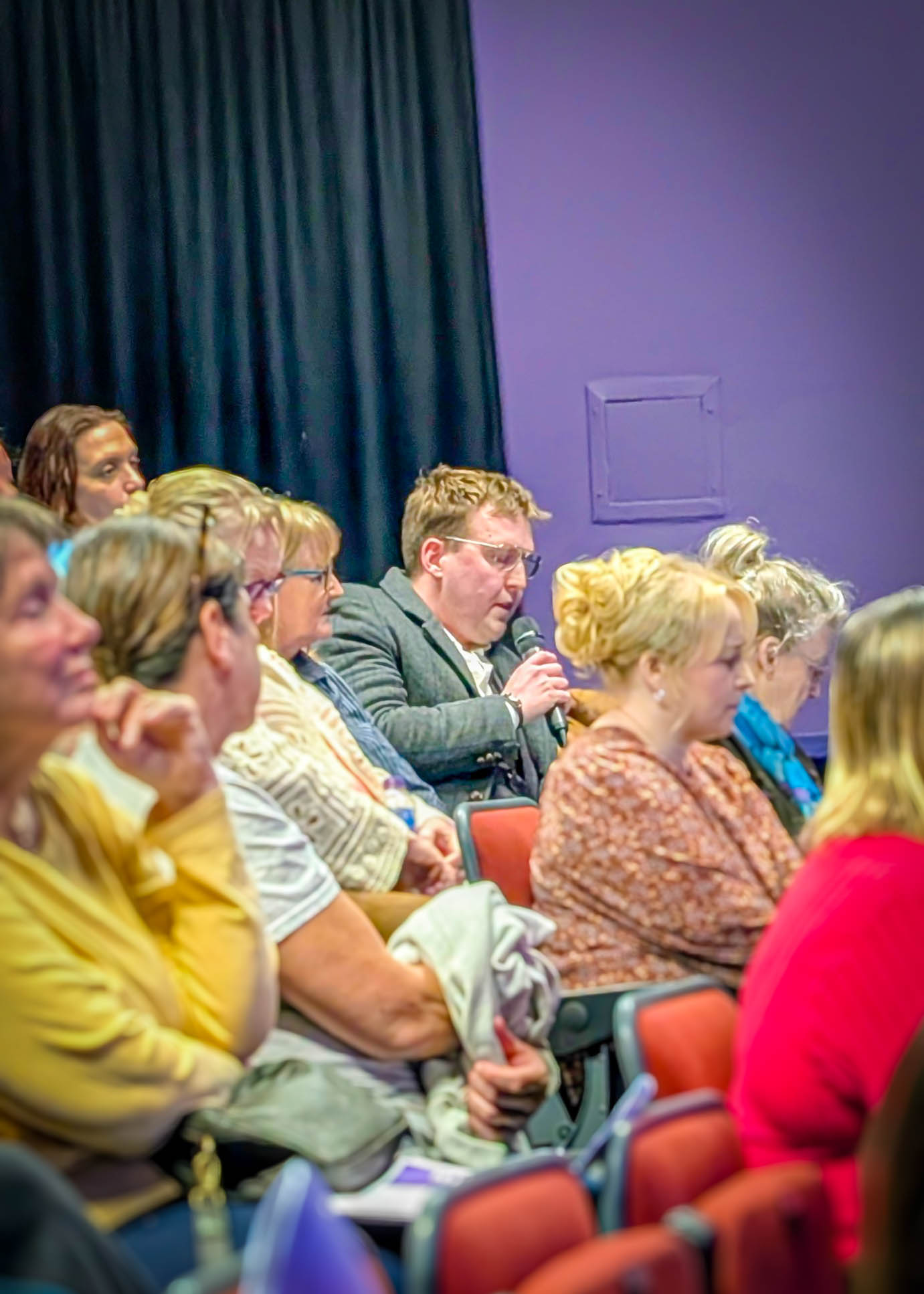 A man in a gray suit holds a microphone and speaks while seated audience members listen in a room with purple walls.