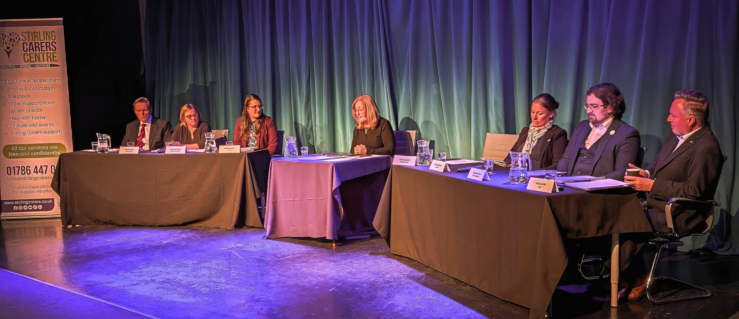 A group of six people sits at two tables on a stage in front of a blue curtain, prepared for a panel discussion.