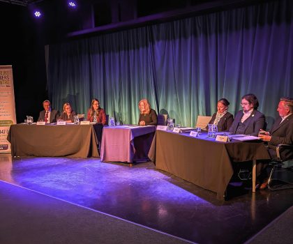 A panel of seven people sits at tables on a stage in front of a blue curtain backdrop.