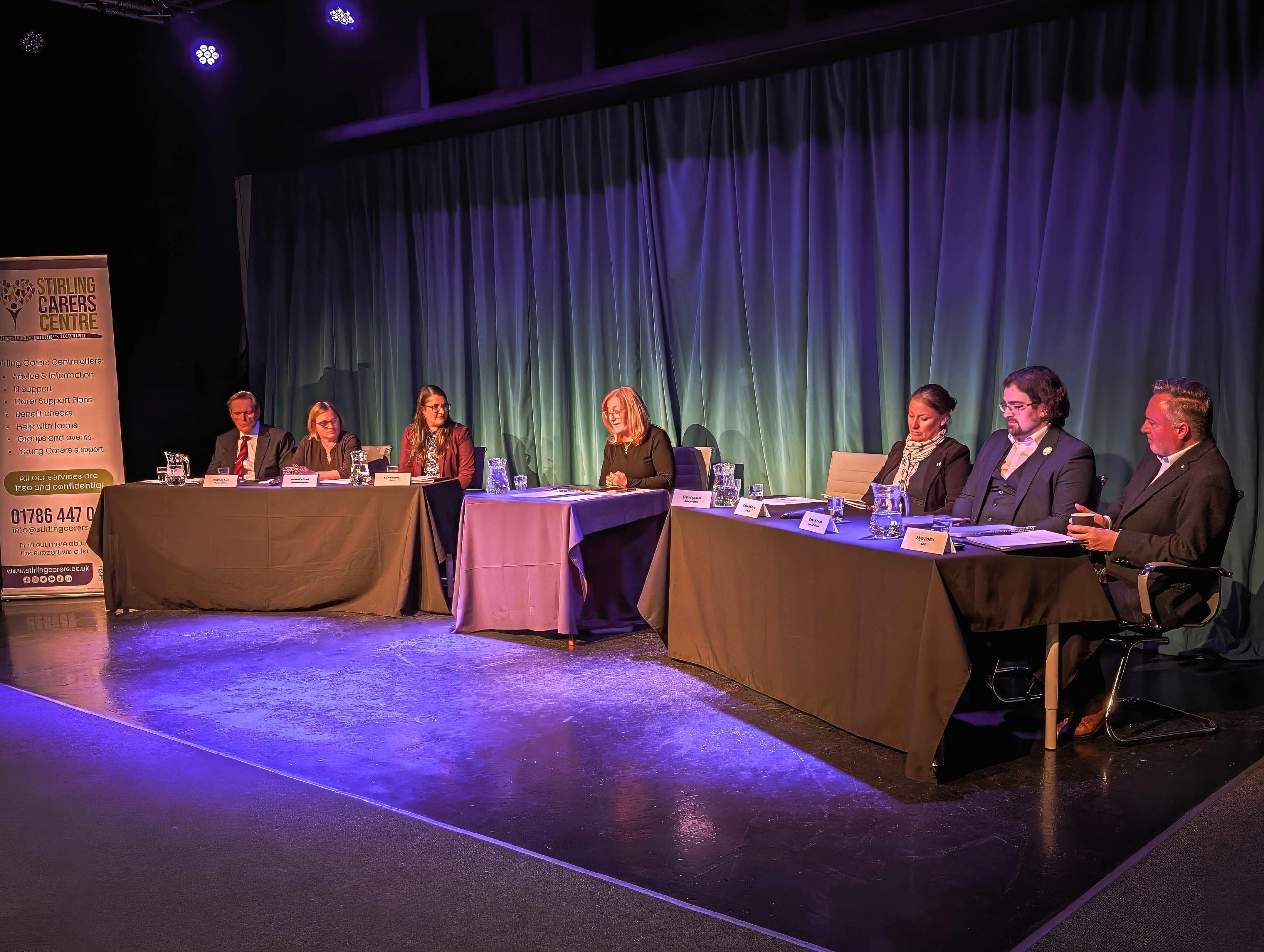 A panel of seven people sits at tables on a stage in front of a blue curtain backdrop.