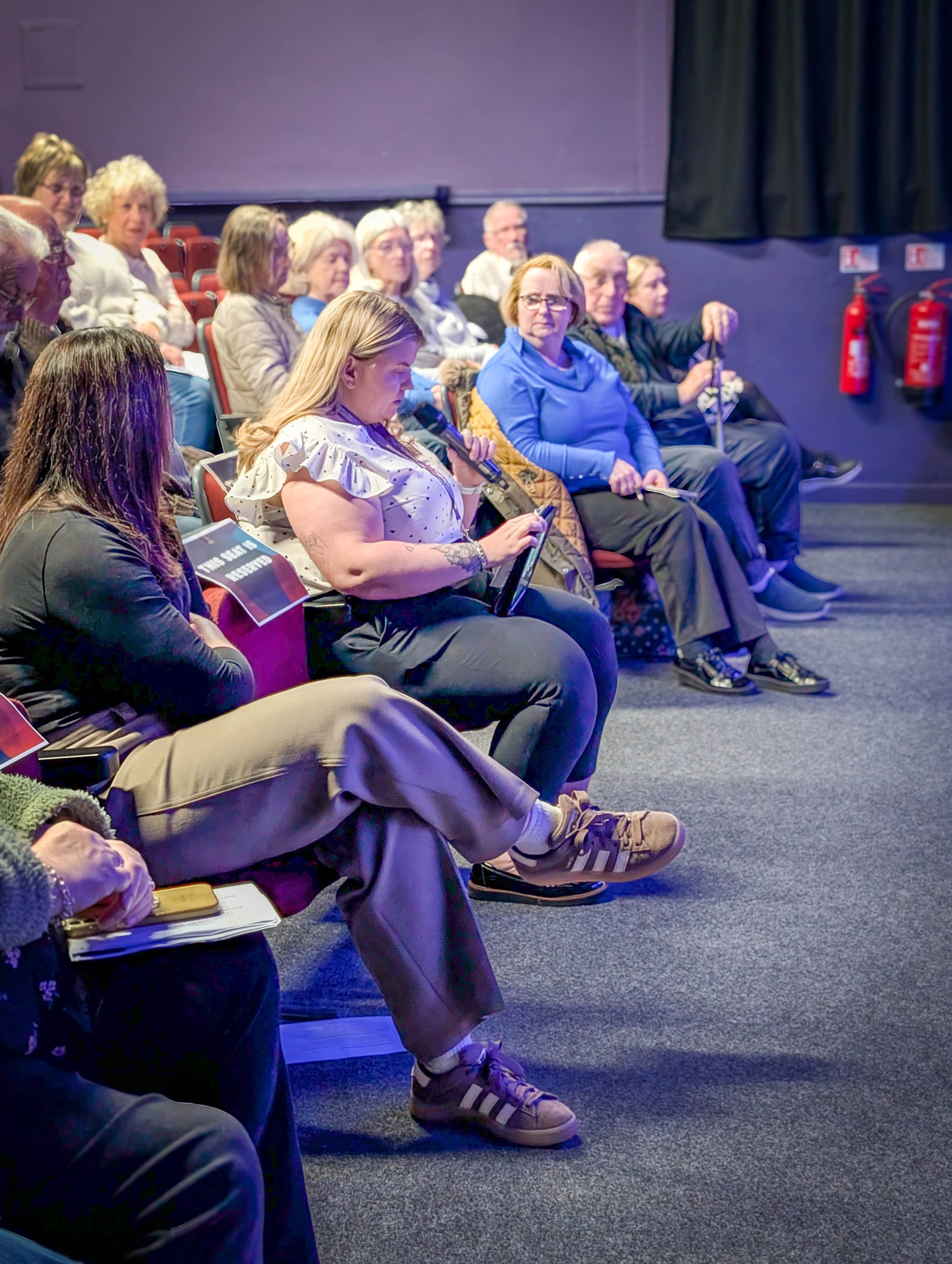 People sit in a well-lit theater; some face forward, while others look at their phones.