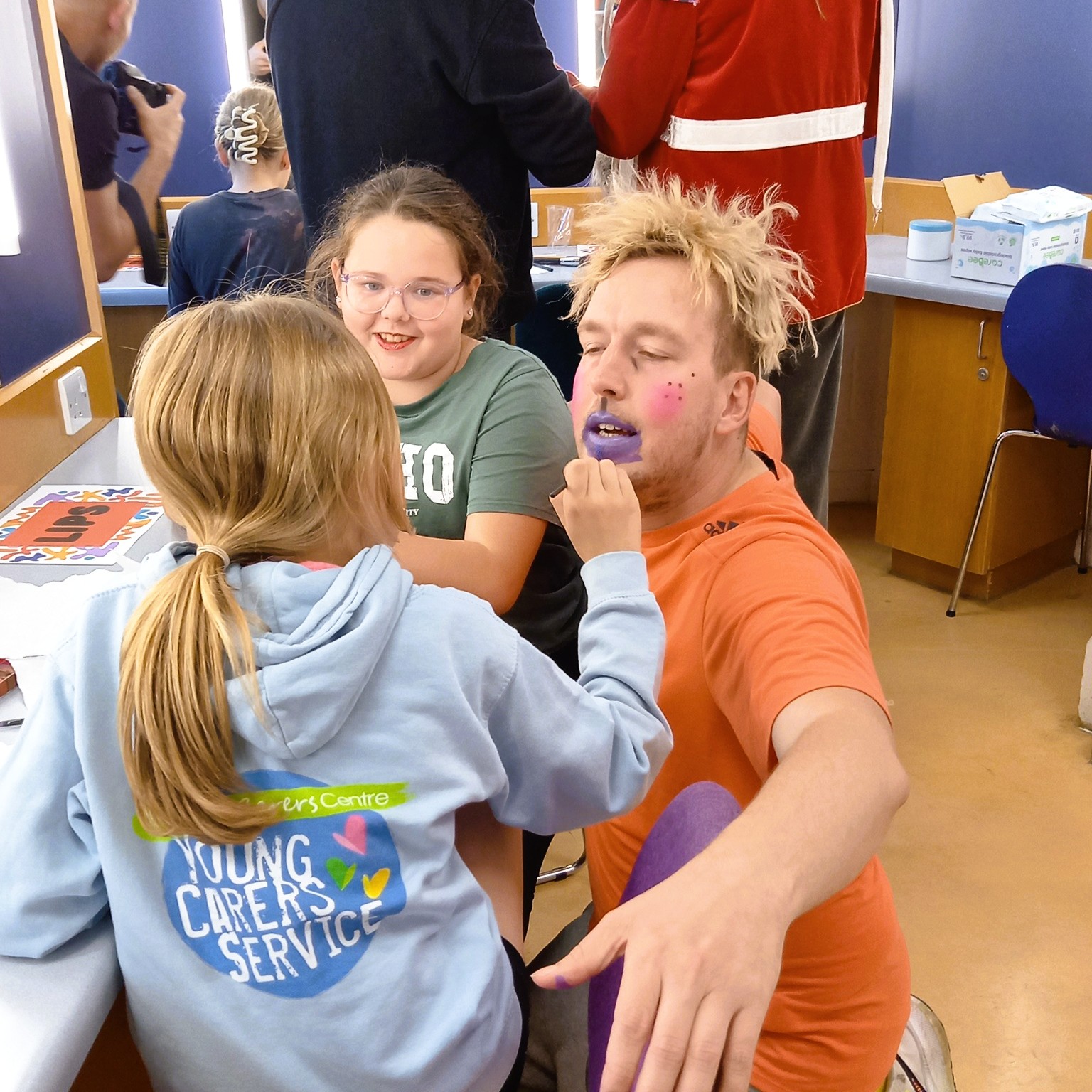 In a lively room, one child applies make-up to a man's face whilst another child stands nearby smiling and observing.