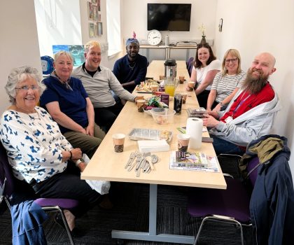 Seven people sit around a table with snacks and drinks, smiling together in a bright, cosy room.