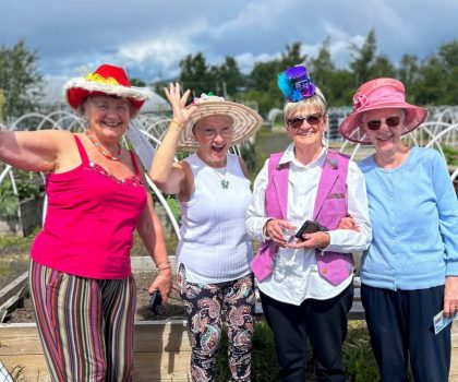 Four older women wearing colourful hats smile and pose together in a sunny outdoor garden.