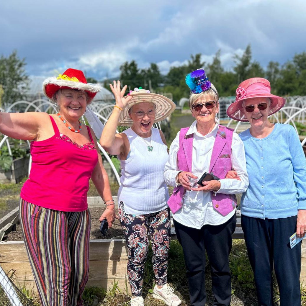Four older women wearing colourful hats smile and pose together in a sunny outdoor garden.