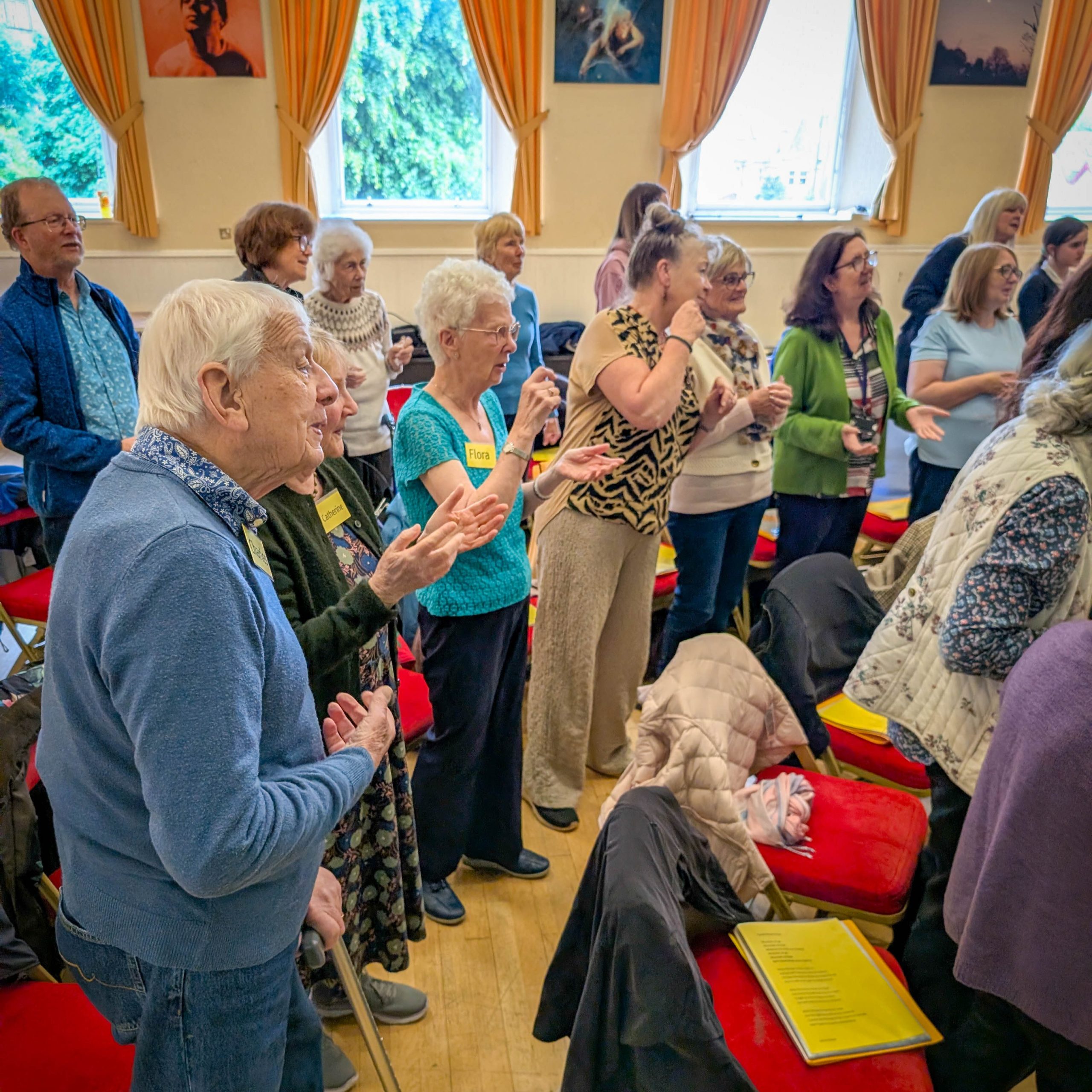 Elderly and middle-aged people stand together and sing in a brightly lit room.