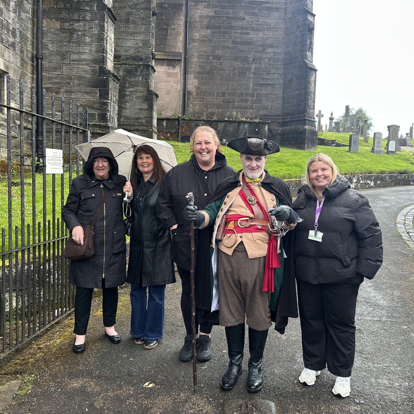 Five people wearing coats, including one in historical costume, stand smiling before a stone building on a rainy day.
