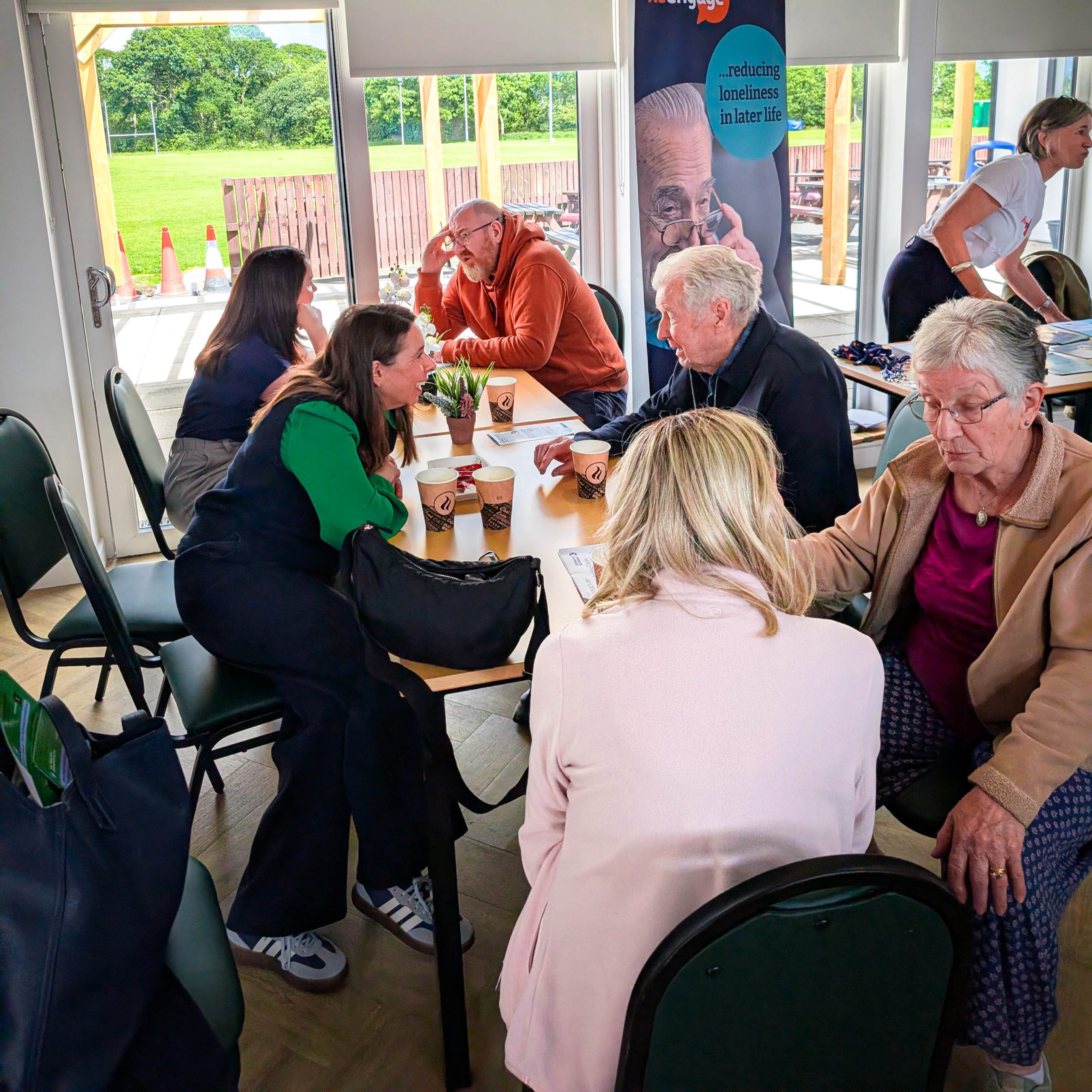 A group of people of different ages sit at tables in a bright room, talking and having coffee, with greenery visible through the windows.