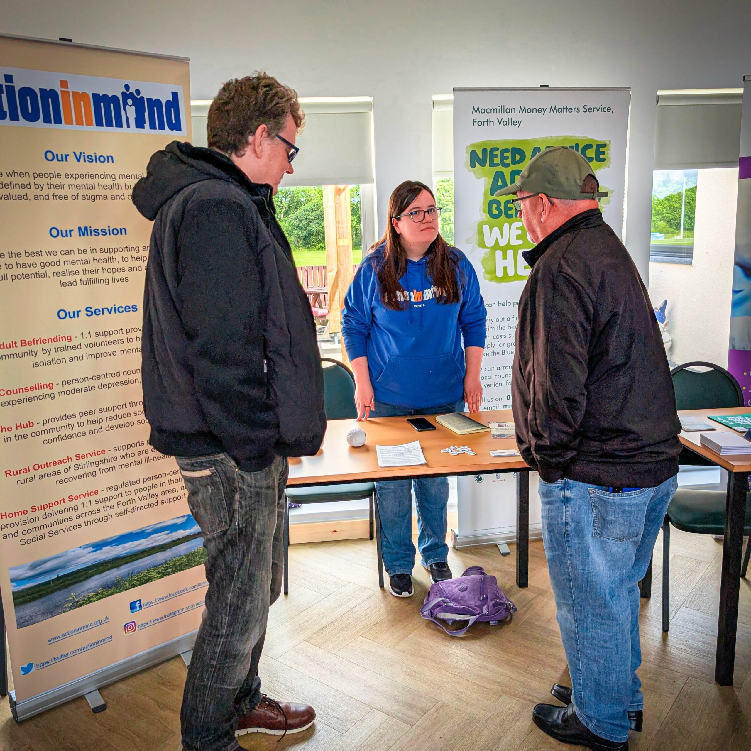 Three people stand and talk at an information stand displaying banners for mental health and money advice services in the background.