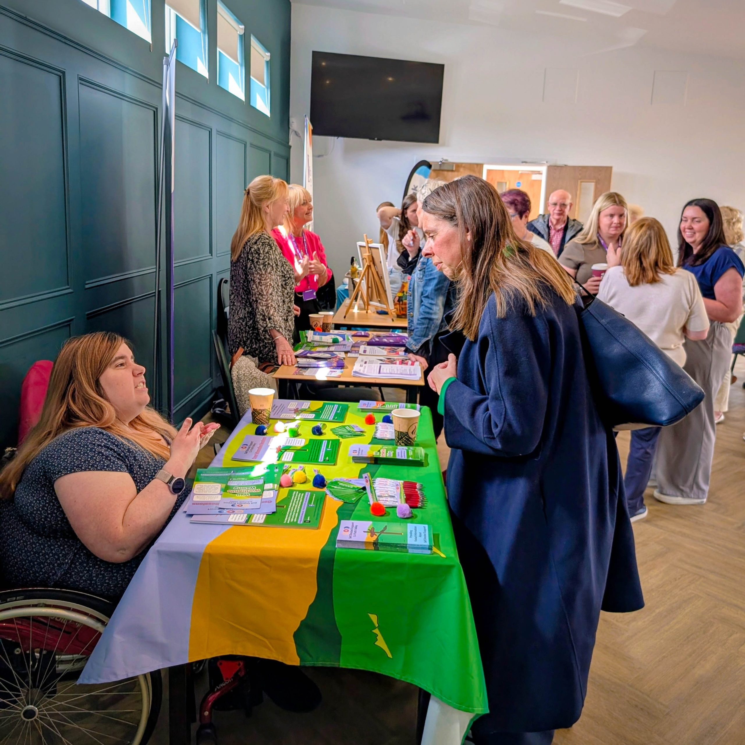 A group of people interact at a stand featuring leaflets and a colourful tablecloth inside an event or fair setting.