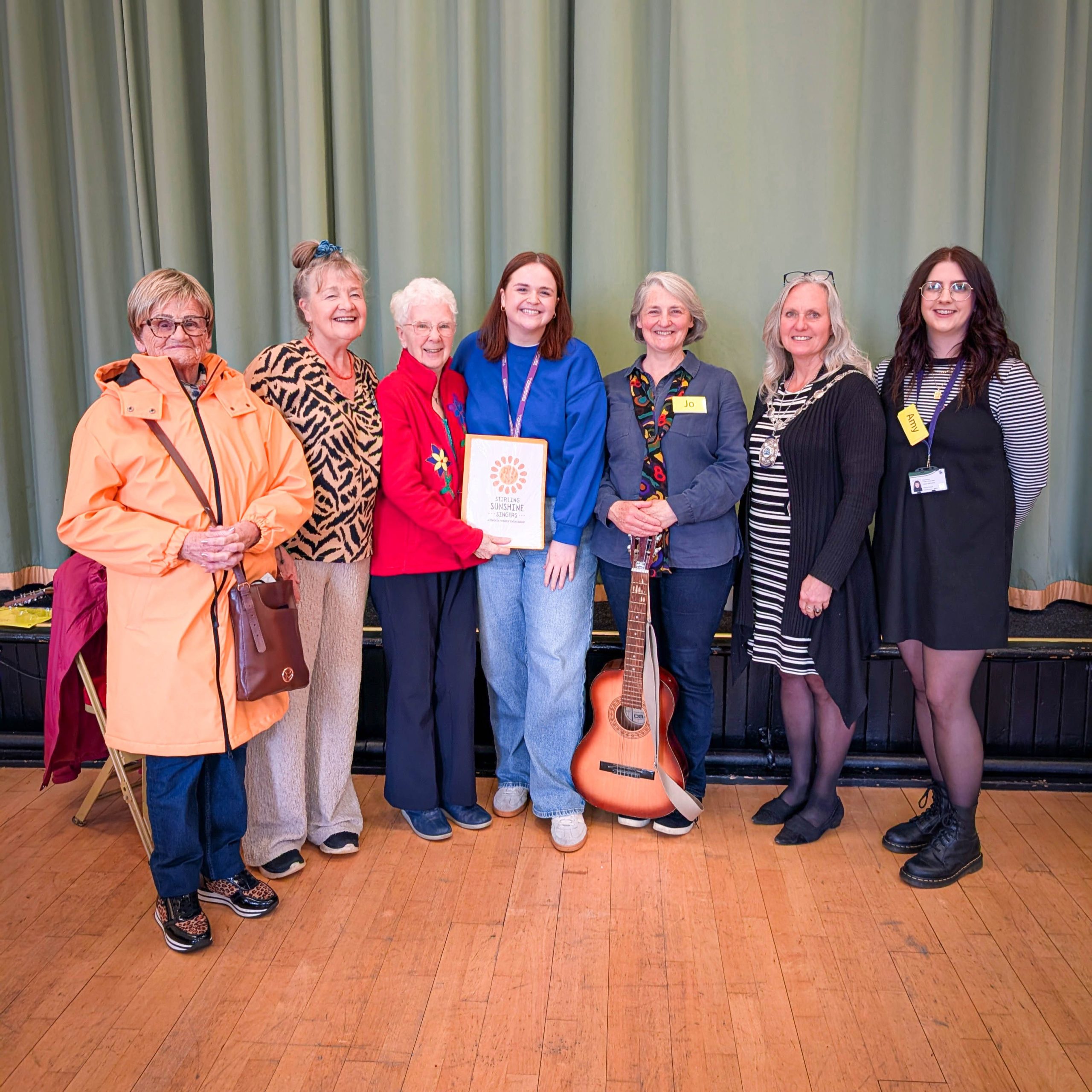 Seven women stand together indoors, smiling, with one holding a certificate and another holding a guitar.