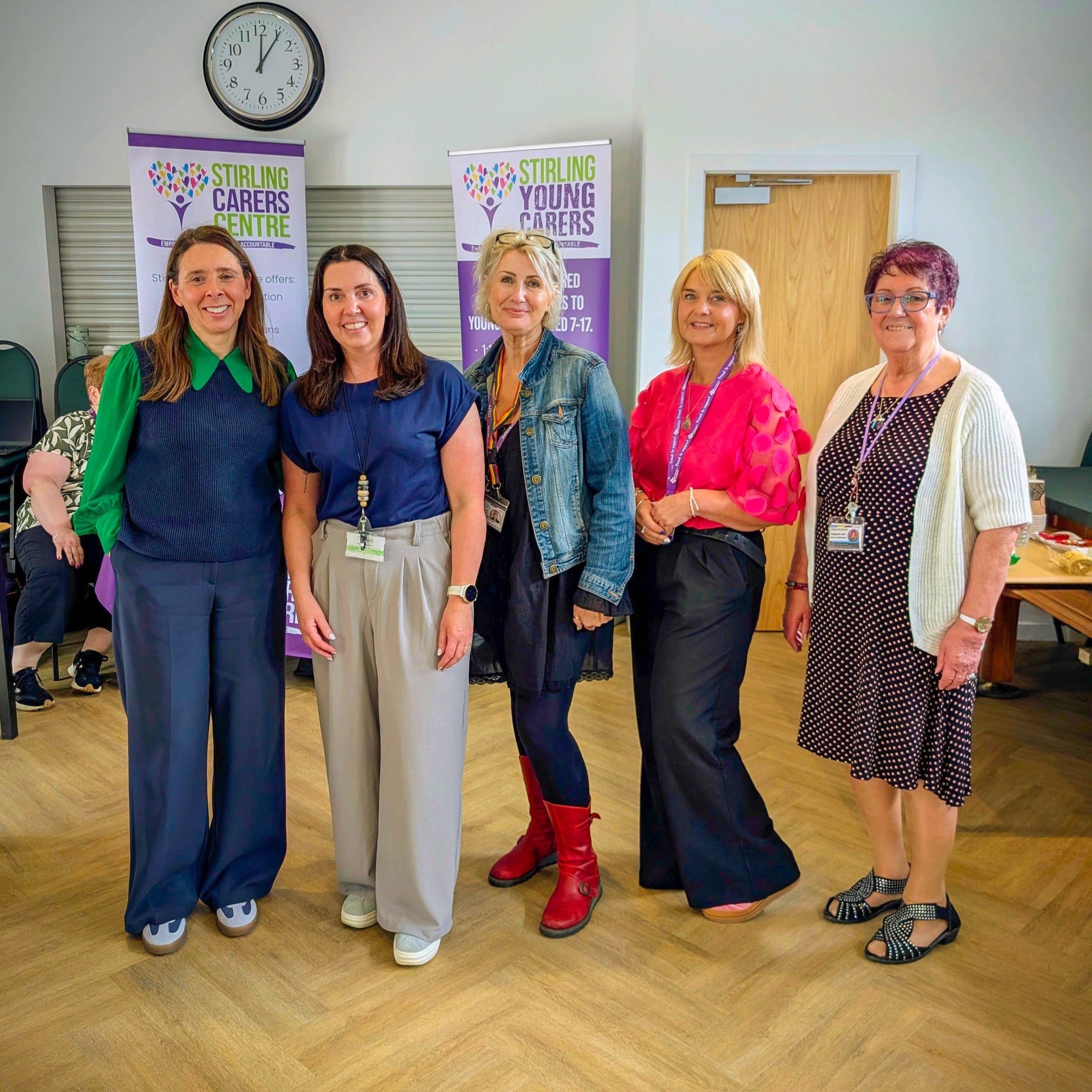 Five women smile whilst standing in a room with banners for Stirling Carers Centre and Stirling Young Carers in the background.