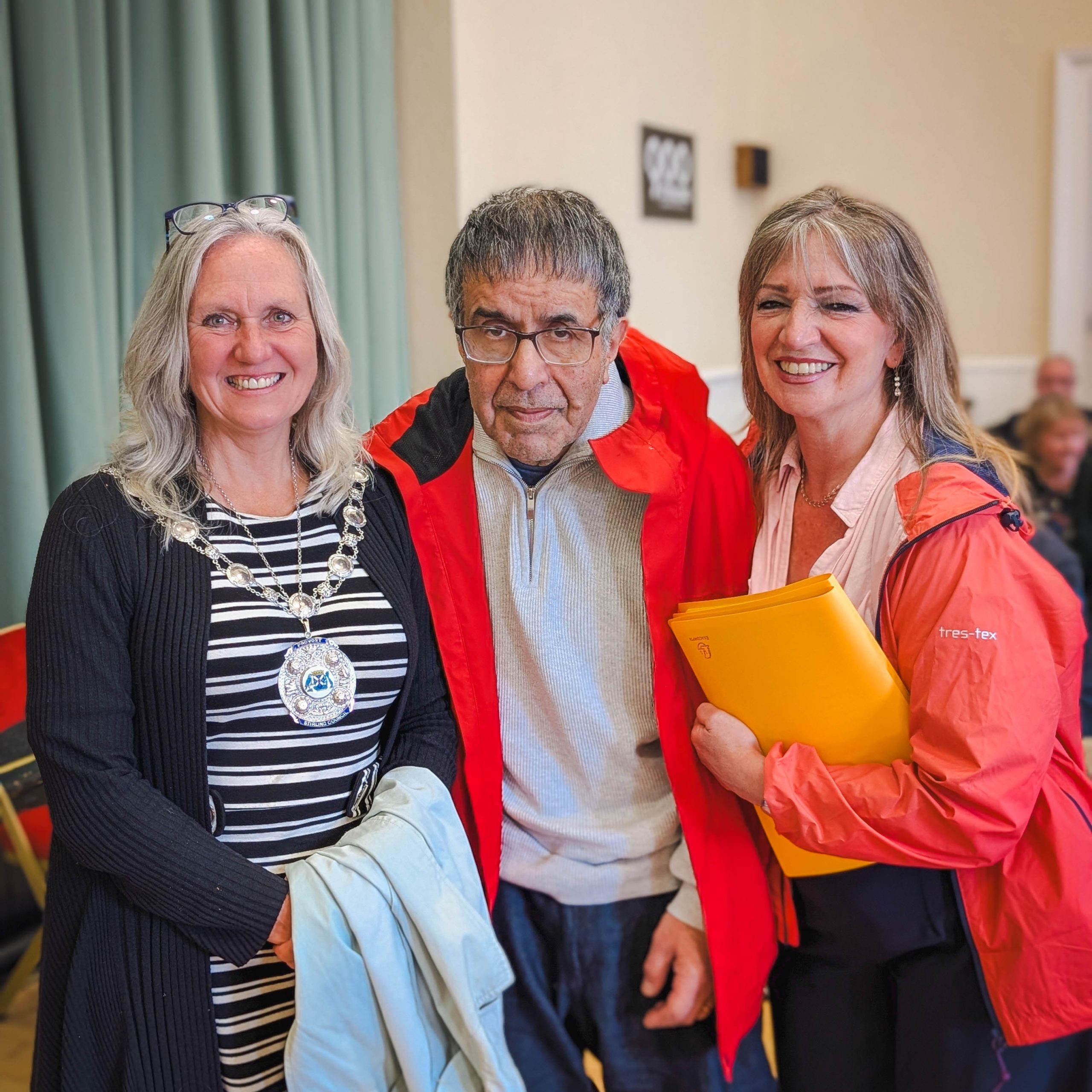 Three adults smile indoors as a man in a red jacket stands between two women, one of whom is wearing a ceremonial chain.