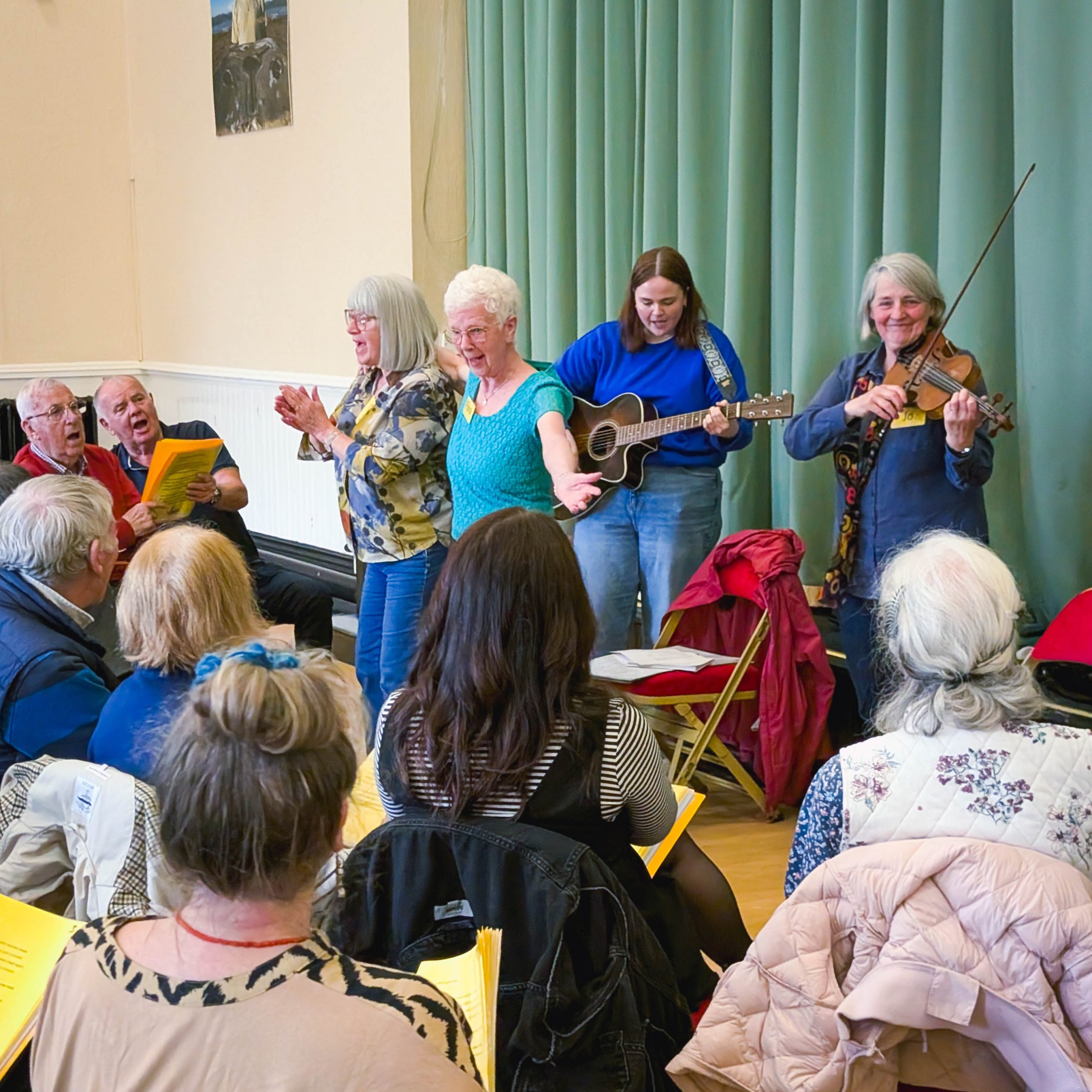Pensioners perform music for an audience inside a community hall decorated with green curtains.