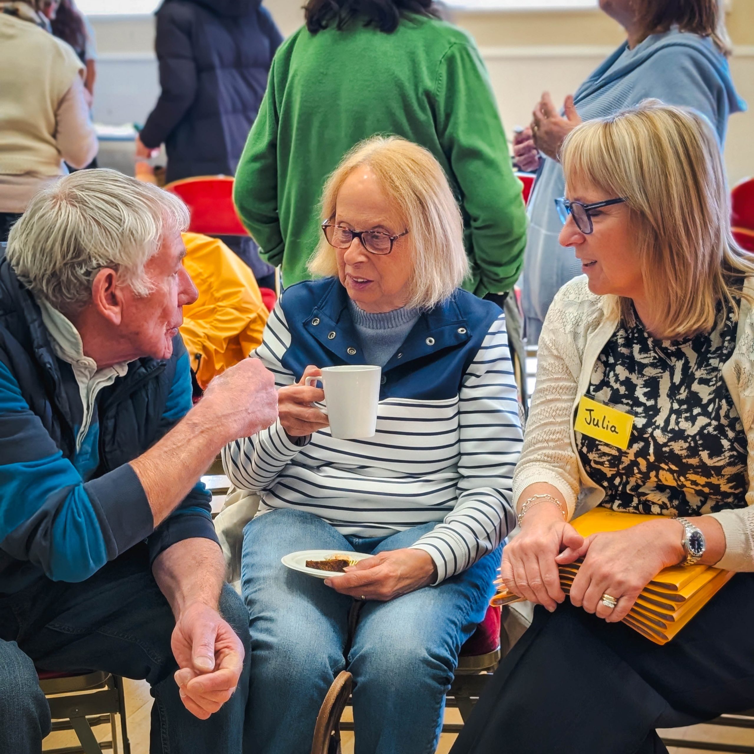 Three adults sit and talk at an indoor gathering; one holds a mug while Julia holds yellow folders.
