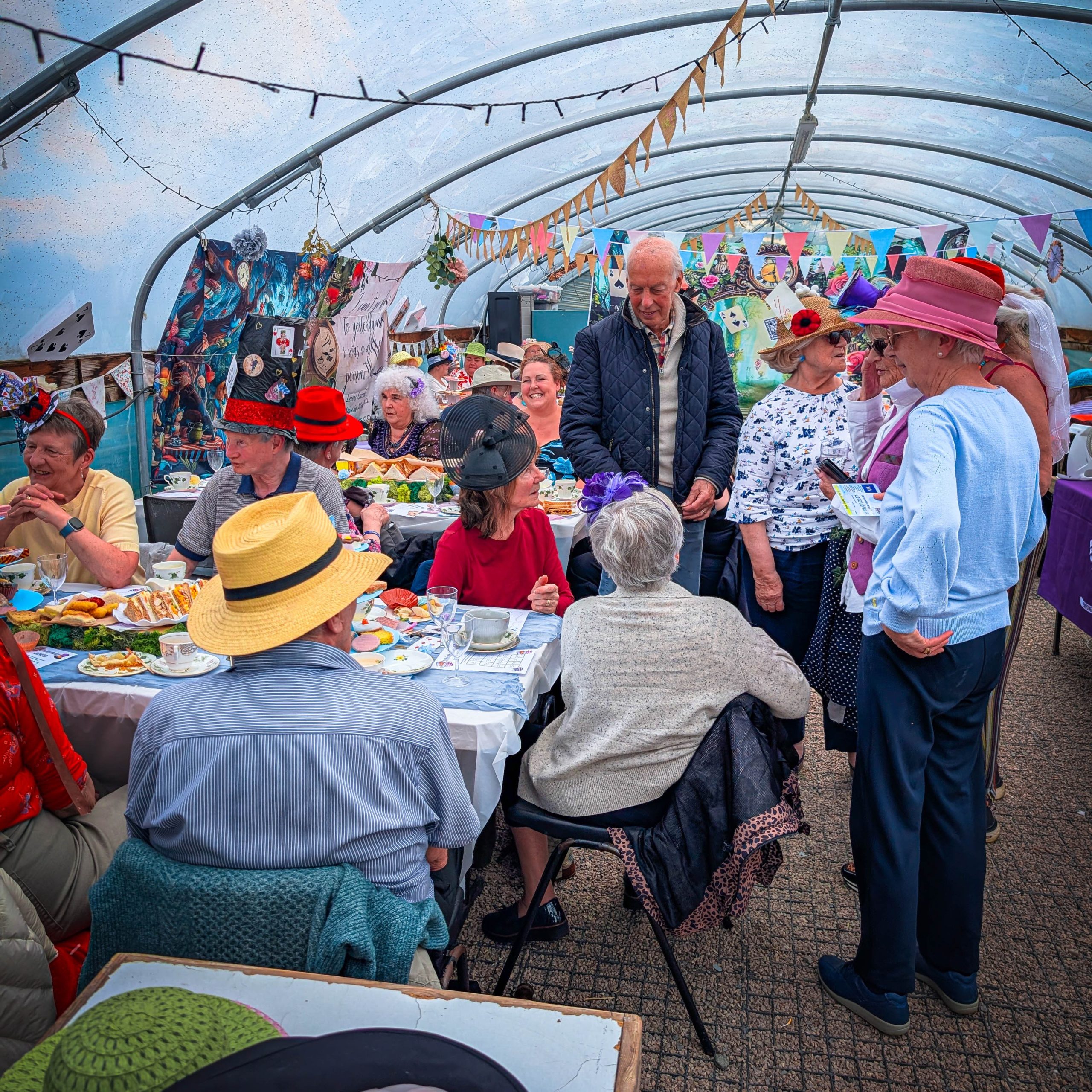 People wearing colourful hats sit together, sipping tea and eating snacks at a vibrant indoor garden party with decorations.