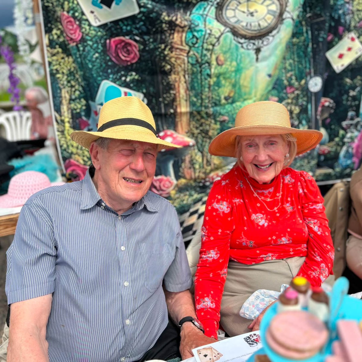 Elderly man and woman wearing straw hats smile together at an outdoor event featuring an Alice in Wonderland themed backdrop.