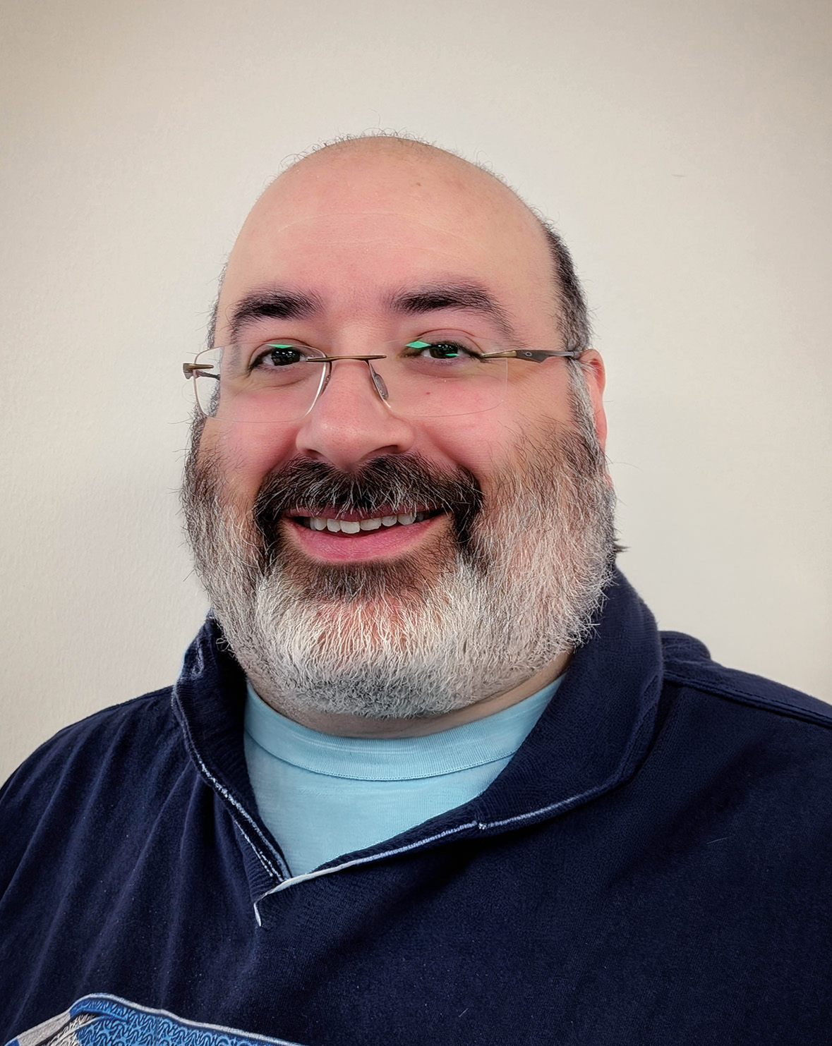A man with glasses, a beard, and moustache, dressed in a navy blue jumper, stands smiling in front of a light background.