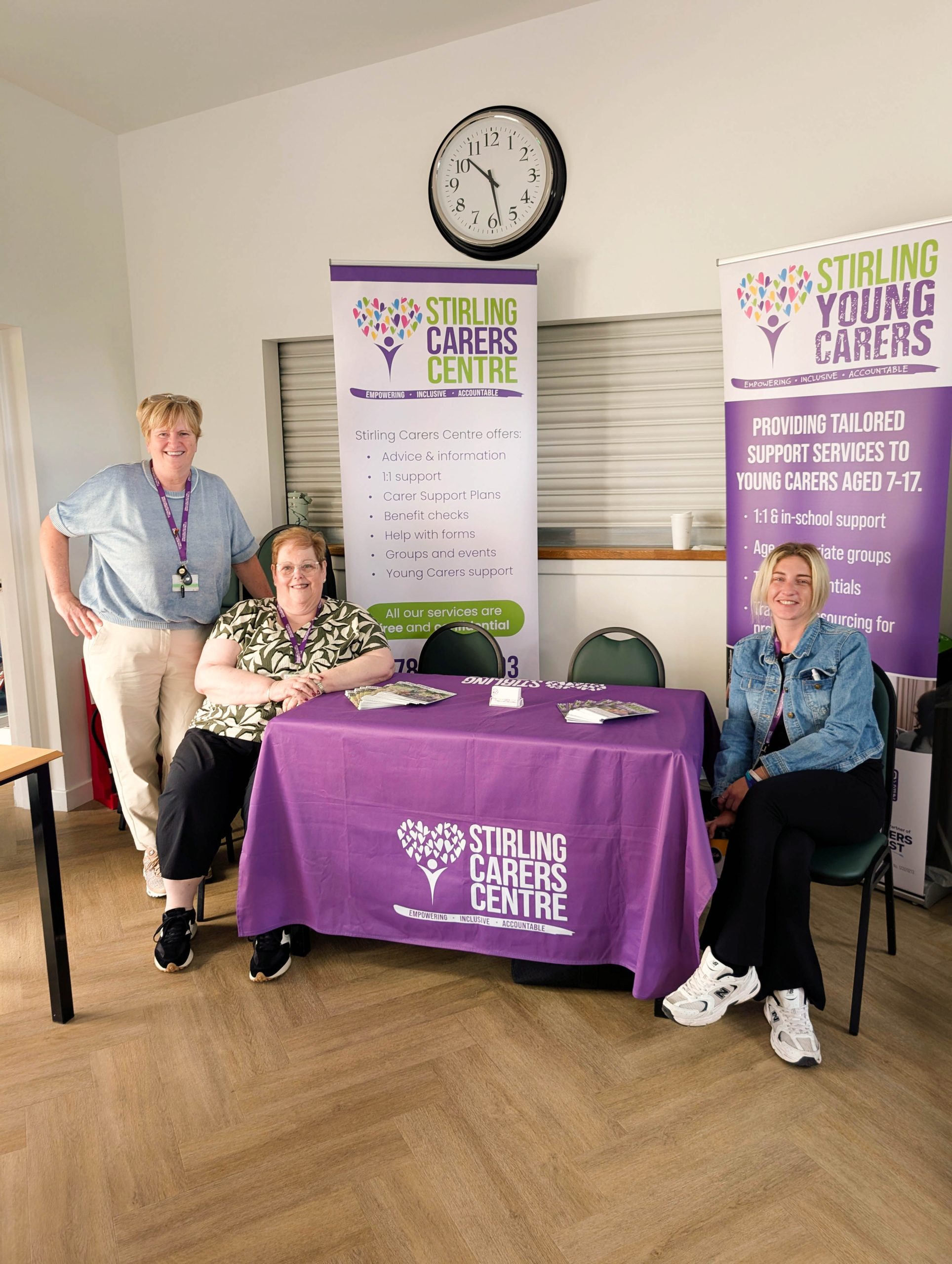 Three people sit behind a Stirling Carers Centre table with informational banners and a purple tablecloth in a well-lit room.