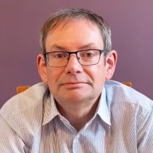 A middle-aged man with short hair and glasses wears a light-coloured striped collared shirt.