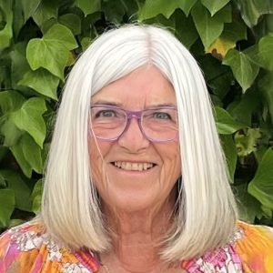 A smiling woman with white hair and purple-rimmed glasses stands in front of ivy leaves.